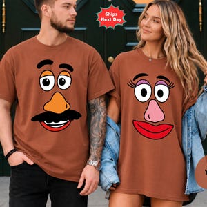 May include: Two brown short-sleeved t-shirts featuring cartoon character faces. The left shirt displays a face with a black mustache, orange nose, and wide eyes. The right shirt features a face with long eyelashes, a pink nose, and red lips.