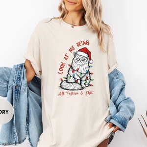 May include: Ivory-colored t-shirt featuring a grumpy cat wearing a Santa hat and Christmas lights. The text reads "LOOK AT ME BEING All Festive & Shit" in red. The shirt is a relaxed fit.