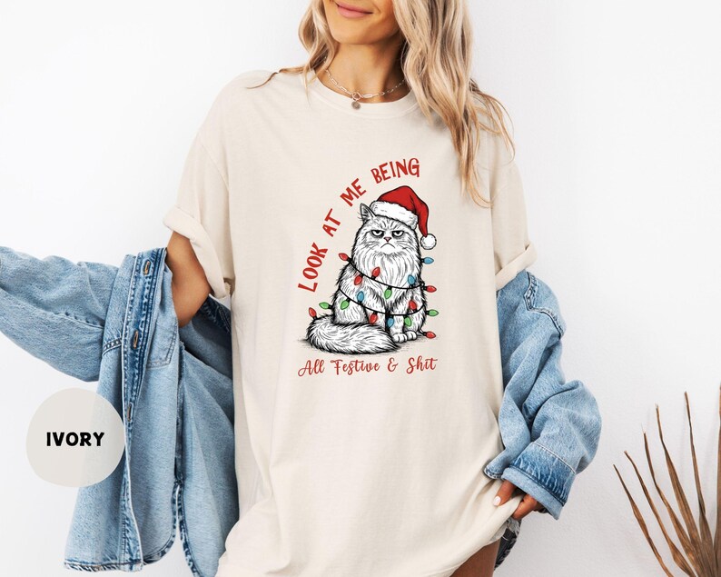 May include: Ivory-colored t-shirt featuring a grumpy cat wearing a Santa hat and Christmas lights. The text reads "LOOK AT ME BEING All Festive & Shit" in red. The shirt is a relaxed fit.