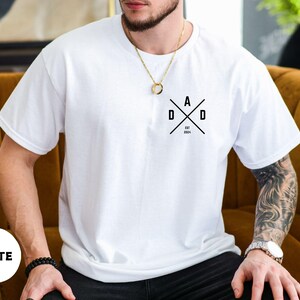 Puede incluir: Camiseta blanca con un dise&ntilde;o gr&aacute;fico negro que dice "DAD" en un dise&ntilde;o cruzado, con "EST 2024" debajo. La camiseta es de color blanco liso y tiene mangas cortas.