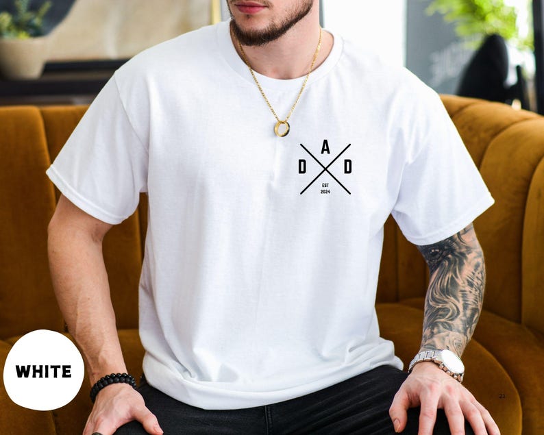 Puede incluir: Camiseta blanca con un dise&ntilde;o gr&aacute;fico negro que dice "DAD" en un dise&ntilde;o cruzado, con "EST 2024" debajo. La camiseta es de color blanco liso y tiene mangas cortas.