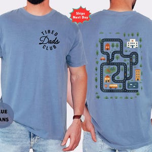 May include: A light blue t-shirt with the text "Tired Dads Club" on the front and a road map design on the back. The back design includes roads, buildings, trees, and vehicles. The shirt is made of a soft material and is suitable for casual wear.