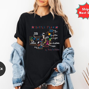 May include: Black t-shirt with a colorful "BATTLE PLAN" graphic. The design includes text and illustrations of various elements like "ice," "fire," and "feathers." The shirt also features the text "by Kevin Mcallister."