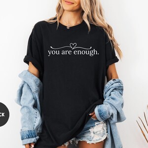 May include: Black t-shirt with the text "you are enough" in a white script font, with a small heart above the word "are". The shirt is paired with a light blue denim jacket and denim shorts. A black circle with the word "BLACK" is in the lower left corner.