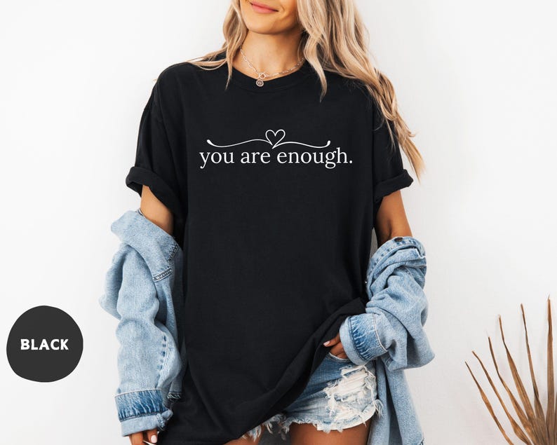 May include: Black t-shirt with the text "you are enough" in a white script font, with a small heart above the word "are". The shirt is paired with a light blue denim jacket and denim shorts. A black circle with the word "BLACK" is in the lower left corner.