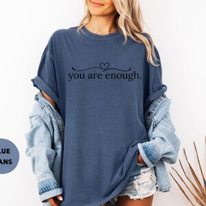 May include: A blue t-shirt with the text "you are enough" in a black script font. The shirt has a relaxed fit and is paired with a denim jacket and shorts. The image also includes a blue circle with the words "BLUE JEANS".