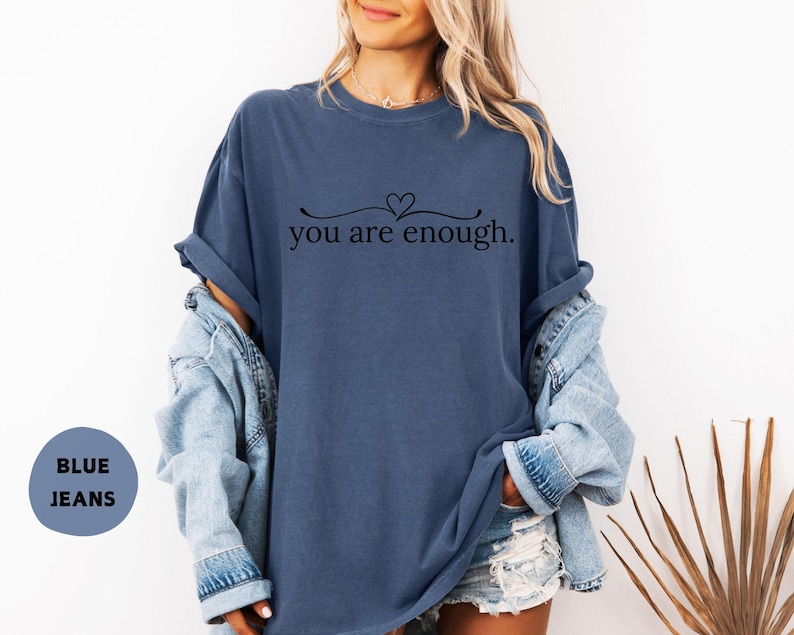 May include: A blue t-shirt with the text "you are enough" in a black script font. The shirt has a relaxed fit and is paired with a denim jacket and shorts. The image also includes a blue circle with the words "BLUE JEANS".