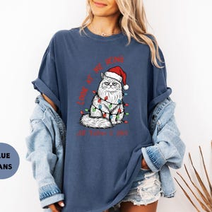 May include: A navy blue t-shirt features a grumpy white cat wearing a Santa hat and Christmas lights. The text "LOOK AT ME BEING... I'M FINE" is printed in red. The shirt is part of a casual wear collection.