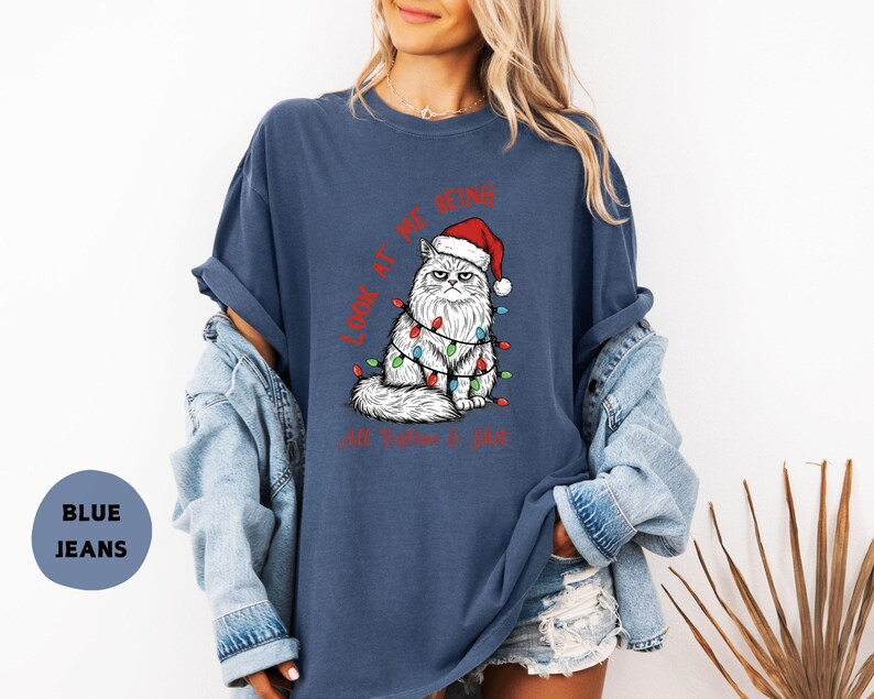 May include: A navy blue t-shirt features a grumpy white cat wearing a Santa hat and Christmas lights. The text "LOOK AT ME BEING... I'M FINE" is printed in red. The shirt is part of a casual wear collection.