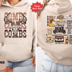 May include: Sand-colored hoodie with a front graphic featuring the name "COMBS" stacked with a deer skull and antlers. The back of the hoodie has various graphics and text, including a cowboy boot, beer bottle, and the phrase "She Got The Best Of Me."
