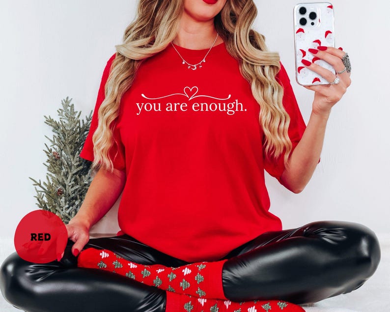 May include: A vibrant red t-shirt featuring the white text "you are enough." with a heart design. The shirt is worn with black leggings and red socks with a green pattern. A phone with a Santa Claus design is held in the hand.