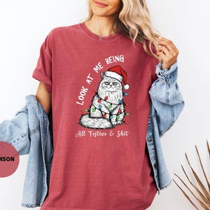 May include: Crimson-colored t-shirt featuring a grumpy cat wearing a Santa hat and Christmas lights. The text "Look at me being All Festive & Shit" is printed on the shirt. The shirt is a comfortable, casual style.