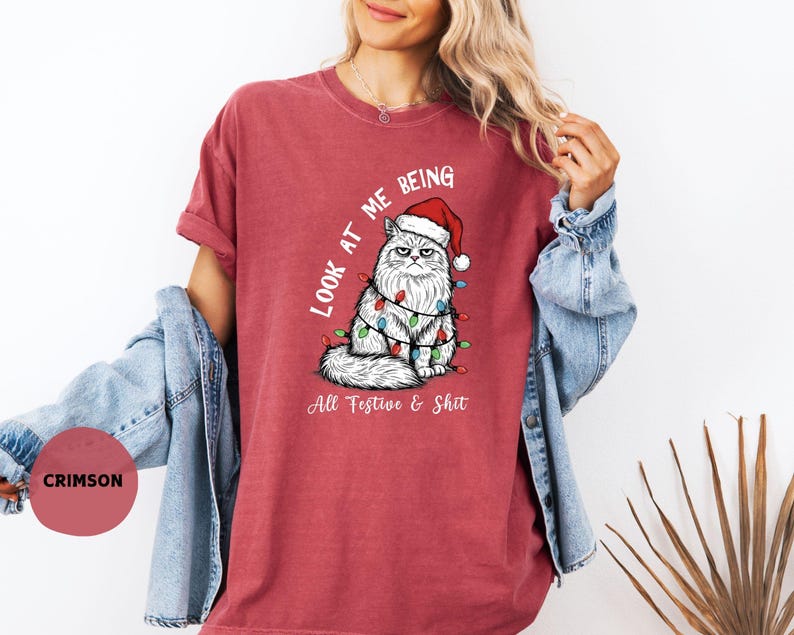 May include: Crimson-colored t-shirt featuring a grumpy cat wearing a Santa hat and Christmas lights. The text "Look at me being All Festive & Shit" is printed on the shirt. The shirt is a comfortable, casual style.