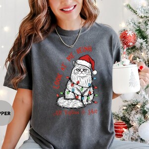 May include: Dark gray t-shirt featuring a grumpy cat wearing a Santa hat and Christmas lights. The text on the shirt reads "LOOK AT ME BEING All Festive Shit". The shirt is a relaxed fit.