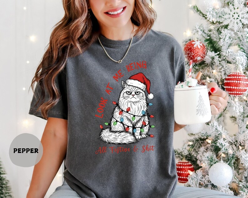 May include: Dark gray t-shirt featuring a grumpy cat wearing a Santa hat and Christmas lights. The text on the shirt reads "LOOK AT ME BEING All Festive Shit". The shirt is a relaxed fit.