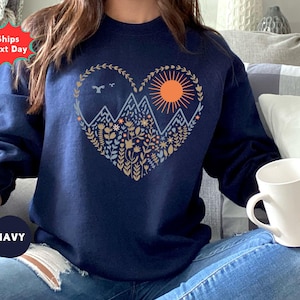 May include: Navy blue sweatshirt featuring a heart-shaped design with mountains, a sun, and floral elements. The design is in shades of beige, orange, and blue. The sweatshirt has long sleeves and a crew neck. The word "NAVY" is printed on a navy blue circle.