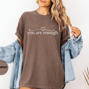 May include: Espresso-coloured t-shirt with the text "you are enough" in a white script. The shirt has a relaxed fit and is paired with a light blue denim jacket. The design includes a heart symbol above the text.