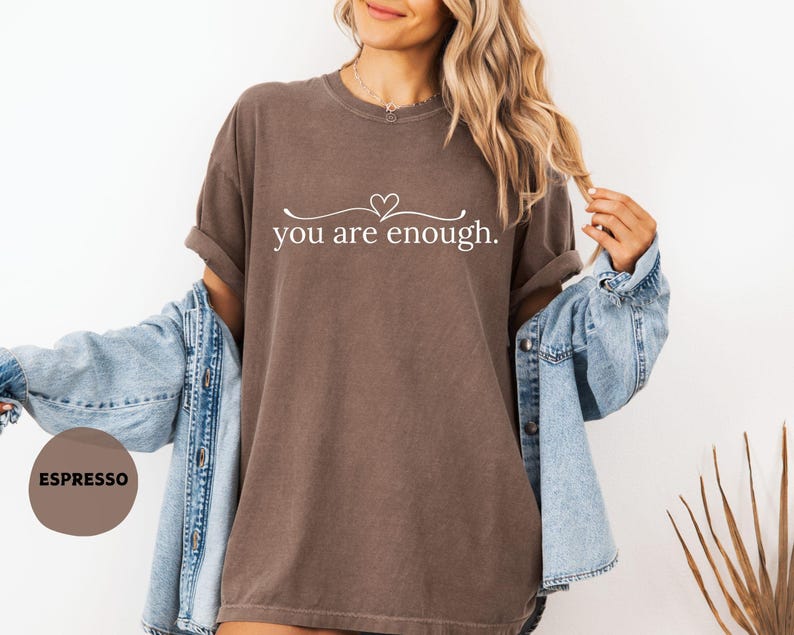 May include: Espresso-coloured t-shirt with the text "you are enough" in a white script. The shirt has a relaxed fit and is paired with a light blue denim jacket. The design includes a heart symbol above the text.