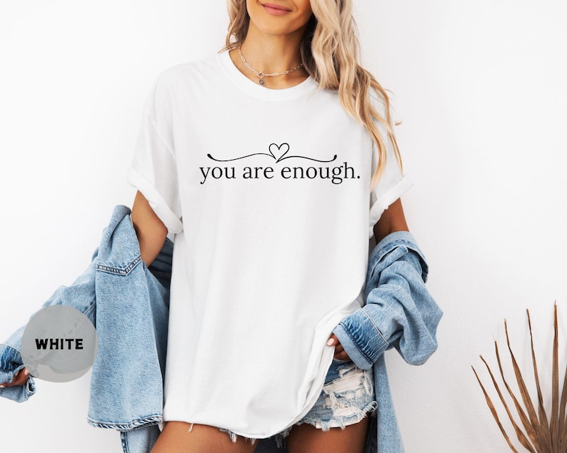 May include: White t-shirt with the text "you are enough" in a black, elegant font. The text is centered and features a heart symbol above the words. The shirt is paired with a denim jacket.