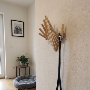 May include: A wooden dog leash holder mounted on a white wall. The holder has multiple wooden dowels arranged in a fan shape. A black dog leash is attached to the holder. The room has hardwood floors, a dog bed, and a framed picture.