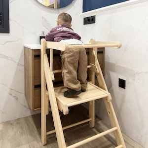 May include: A natural wood learning tower with a step-stool design. The tower has a platform and a safety rail. The tower is designed to help children reach countertops and sinks. The tower is in a bathroom setting.