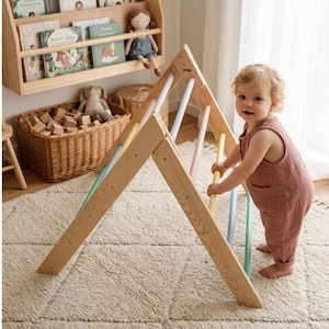 May include: A triangular wooden climbing frame featuring light-colored wood side panels and a series of horizontal rungs in pastel shades of yellow, pink, and light blue. The structure is designed for indoor play and has a sturdy, open frame.