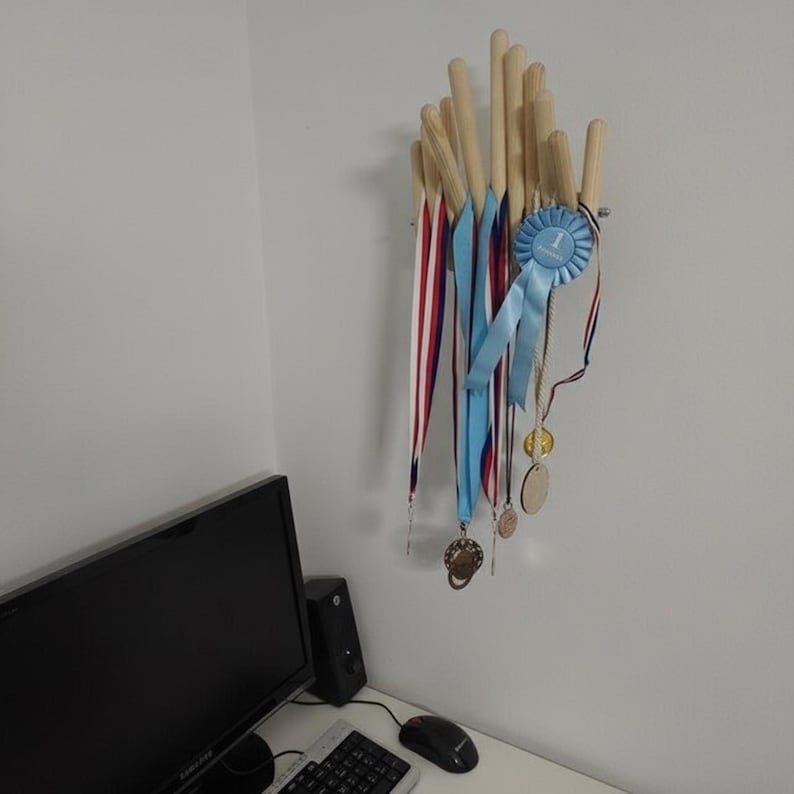 May include: A wooden medal display rack with multiple dowels. Ribbons in red, white, and blue, along with a blue ribbon and medals, hang from the rack. The rack is mounted on a white wall.