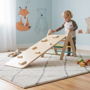 Puede incluir: Un juego de arco y tobogán de escalada de madera con un acabado de color claro. El tobogán tiene bloques de madera adjuntos para escalar. El juego está sobre una alfombra estampada, con un niño interactuando con el juguete. Se ve arte mural de un zorro y un oso.