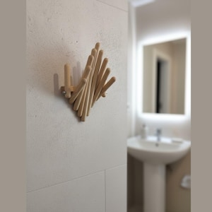 May include: A wooden wall-mounted coat rack with a geometric design. The rack is made of multiple light-colored wooden dowels arranged in a diamond shape. It is mounted on a white tiled wall, suitable for a bathroom or entryway.