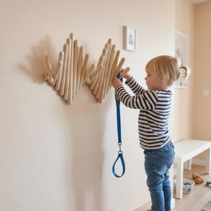 Könnte beinhalten: Eine Wandgarderobe aus hellem Holz mit einer Reihe vertikaler, verstellbarer Stäbe, die in einem wellenförmigen Muster angeordnet sind. Die Garderobe verfügt über mehrere Haken zum Aufhängen von Gegenständen, an denen eine blaue Leine hängt.