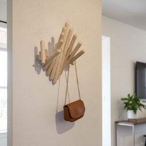 Könnte beinhalten: Ein hellfarbiger Wandgarderobenständer aus Holz mit mehreren Haken in einem einzigartigen, künstlerischen Design. Eine braune Ledertasche mit goldener Kette hängt an einem der Haken. Der Ständer ist an einer neutralfarbenen Wand befestigt.