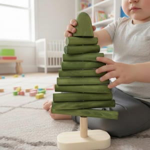 Può includere: Un giocattolo in legno verde a forma di albero, con base e tronco in legno chiaro. L'albero è composto da pezzi cilindrici impilati. Un bambino tiene in mano il pezzo superiore dell'albero. Il giocattolo è su un tappeto chiaro.