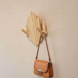 May include: A light-colored wooden wall-mounted coat rack with a geometric design. A brown leather purse with a silver clasp hangs from one of the pegs. The rack is mounted on a beige wall.