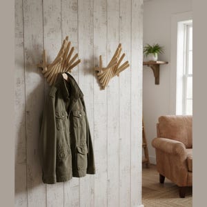May include: Two wooden wall-mounted coat racks with a unique, geometric design. One rack holds an olive green jacket. The racks are mounted on a white, textured wall. A small shelf with a plant is visible in the background.
