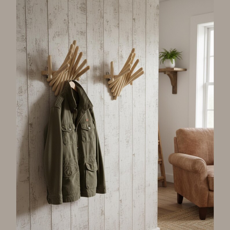 May include: Two wooden wall-mounted coat racks with a unique, geometric design. One rack holds an olive green jacket. The racks are mounted on a white, textured wall. A small shelf with a plant is visible in the background.