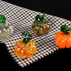 Puede incluir: Cuatro calabazas de vidrio con tallos verdes descansan sobre una tela a cuadros. Una es transparente, otra naranja y dos son multicolores con motas rojas, amarillas y azules. Las calabazas son una decoración festiva de otoño.