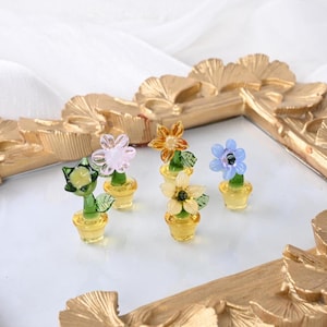Könnte beinhalten: Eine Sammlung von Miniatur-Glasblumenskulpturen in verschiedenen Farben, darunter Rosa, Orange, Gelb und Blau. Jede Blume sitzt in einem kleinen gelben Topf. Die Blumen sind auf einem goldfarbenen dekorativen Rahmen angeordnet.