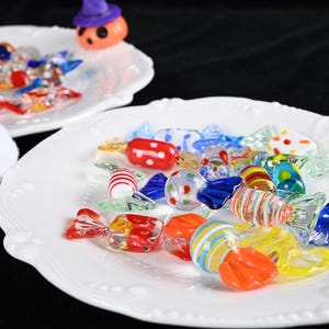 Puede incluir: Un plato blanco lleno de caramelos de cristal de colores en varias formas y diseños. Los caramelos están envueltos para parecer caramelos envueltos. Una pequeña figura de calabaza con un sombrero morado está en el fondo.