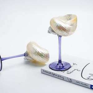 May include: Two decorative wine glasses with iridescent seashell bowls and purple stems and bases. One glass is upright, the other is lying on its side. A book with the title "The Unbearable Lightness of Being" is in the image.