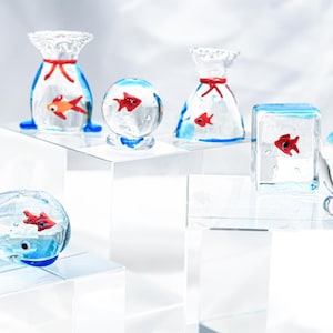 Puede incluir: Colección de esculturas de vidrio con peces dorados. Las esculturas tienen varias formas, incluyendo una ballena, esferas y bolsas. Cada pieza tiene un pez dorado rojo en su interior, con detalles azules y blancos.