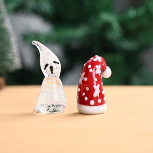 以下が含まれることがあります： 2つのガラス製のクリスマスオーナメント：黒い顔の特徴を持つ透明な幽霊と、白い雪の結晶のデザインと白いポンポンが付いた赤いサンタの帽子。 オーナメントは、ぼやけた緑色の背景を持つ明るい茶色の表面にあります。