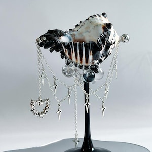 以下が含まれることがあります： 装飾的な貝殻のアート作品。貝殻は黒と白で、黒いスタンドに取り付けられています。星とハートのチャームが付いたシルバーのチェーンが貝殻から吊り下げられ、透明なガラスビーズが付いています。ベースは反射性です。
