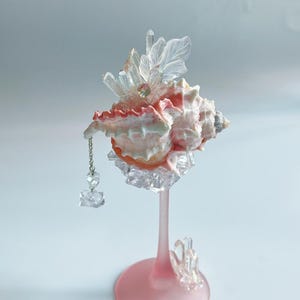 May include: A decorative seashell sculpture. The shell is primarily pink and white, adorned with clear crystal accents and a dangling chain with a crystal pendant. It is mounted on a pink stem and base, with additional crystal embellishments.