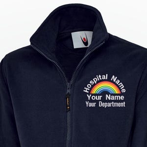 Könnte beinhalten: Marineblaue Fleecejacke mit durchgehendem Reißverschluss und hohem Kragen. Die Jacke hat eine Regenbogen-Grafik und den Text "Hospital Name, Your Name, Your Department" in Weiß. Zwei Vordertaschen sind vorhanden.