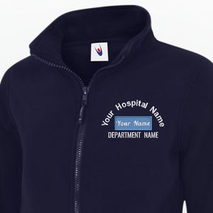 Può includere: Giacca in pile blu navy con zip e collo alto. La giacca ha due tasche anteriori e un piccolo logo sulla parte superiore sinistra del petto. Il testo sul petto recita "Your Hospital Name", "Your Name" e "DEPARTMENT NAME".