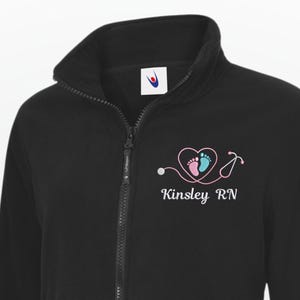 Könnte beinhalten: Schwarze Fleecejacke mit Reißverschluss und hohem Kragen. Die Jacke hat ein gesticktes Design eines Herzens mit Babyfüßen und einem Stethoskop sowie den Text "Kinsley RN". Die Jacke hat zwei Vordertaschen.
