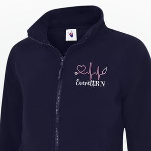 May include: Navy blue fleece zip-up jacket with a high collar. Features a pink heartbeat design and the text "Everett RN". The jacket has two front pockets and a full-length zipper.