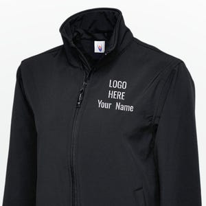 Op de afbeelding: Zwarte jas met een ritssluiting over de hele lengte, een opstaande kraag en twee zijzakken. De tekst "LOGO HERE Your Name" is in het wit gedrukt op de linkerbovenborst. De jas is gemaakt van een glad, mat materiaal.