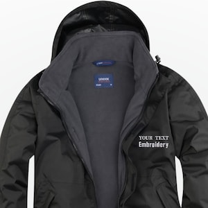 Puede incluir: Chaqueta negra con capucha y forro interior gris. La chaqueta tiene cierre de cremallera y el texto "YOUR TEXT Embroidery" es visible en el pecho derecho. Diseñada para uso en exteriores.