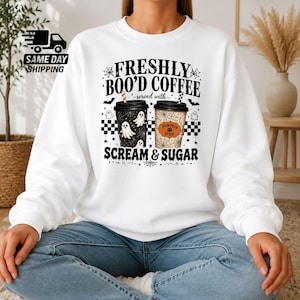 May include: A white crewneck sweatshirt featuring a graphic of two coffee cups with the text FRESHLY BOO'D COFFEE served with SCREAM & SUGAR. The design includes ghost illustrations, spiderwebs, and a pumpkin spice label on a checkered background.